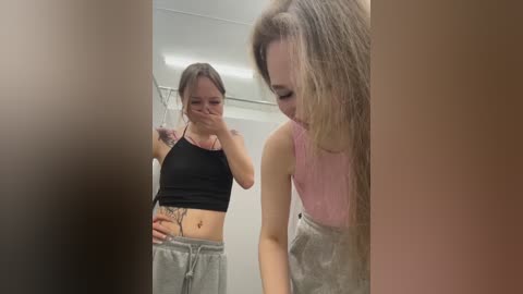 Video of a young woman with long blonde hair, wearing a pink top and gray sweatpants, covering her face with her hand in a dimly lit bathroom.