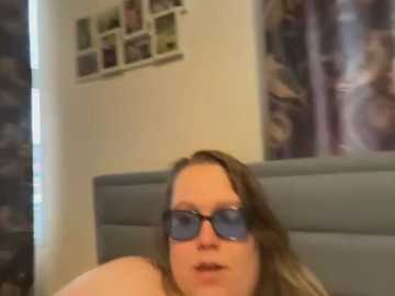 A blurry video of a woman with glasses and brown hair, wearing a light-colored shirt, sitting on a bed with a grey headboard. The background includes a collage of photos and a purple curtain.