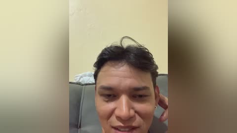 Video of a man with short black hair and a small smile, sitting on a grey couch in a plain room with beige walls.