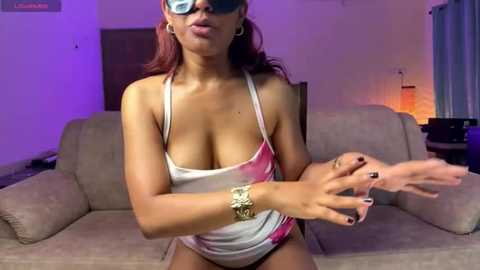 Video of a curvy, dark-skinned woman with large breasts, wearing a pink-and-white tie-dye tank top, blue sunglasses, and a gold bracelet, posing on a beige couch in a dimly lit room.