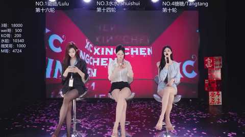 Video of three Asian women in revealing outfits, seated on stools on a stage with a red and pink backdrop, holding microphones.