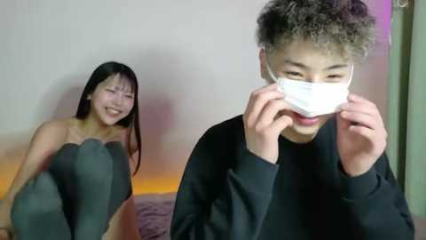 Video of a smiling Asian woman in a black tank top, seated on a bed, and a young Asian man in a black shirt, covering his face with a white mask.