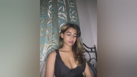 Video of a young woman with medium brown skin, wearing a low-cut black lace dress, standing in a room with teal and white patterned curtains and a black wrought iron bed.