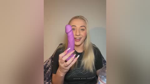 Video of a blonde woman with fair skin, wearing a black graphic tee, holding a purple dildo, smiling in a playful manner.
