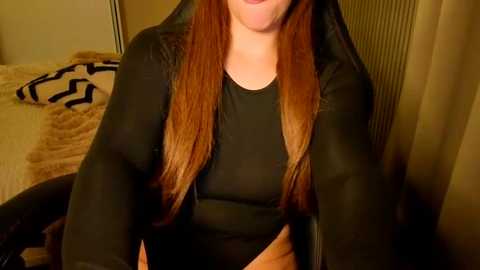 Video of a woman with long, straight, light brown hair, wearing a black long-sleeve shirt. She sits on a bed with a beige comforter and a black and white patterned pillow. The room has beige walls and a radiator.