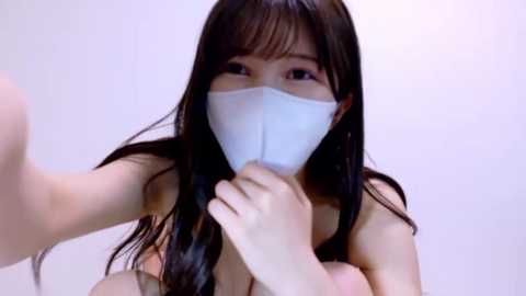 Video of an East Asian woman with long black hair, wearing a white face mask, holding a mask to her mouth, revealing bare shoulders, against a plain white background.