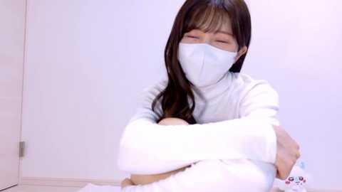 Video of an East Asian woman with long, straight dark hair, wearing a white face mask, light-colored turtleneck sweater, and white pants, sitting cross-legged on a white floor with a teddy bear.