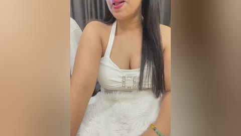 Video of a Latina woman with medium brown skin, long dark hair, and full lips, wearing a white lace dress with a plunging neckline, revealing ample cleavage. She stands against a beige wall, smiling slightly.