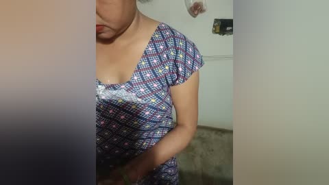 Video of a woman with light brown skin, wearing a patterned blue dress, standing in a dimly lit room with a beige carpet.