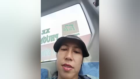 A video of a young woman with light skin, wearing a black hat, blue shirt, and blue seatbelt, smiling, taken inside a car with a blurred storefront in the background.