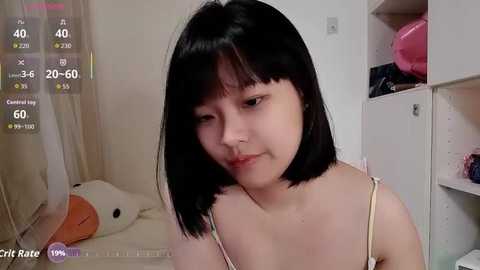 Video of a young Asian woman with straight black hair, light skin, wearing a beige spaghetti-strap top, sitting in a bedroom with white walls and a stuffed toy.