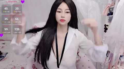 A video of an East Asian woman with long black hair, wearing a white shirt with a deep V-neck, posing in a cluttered room.