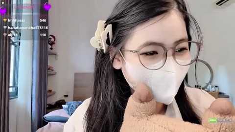 A video shows a young Asian woman with long black hair and glasses, wearing a mask, being kissed passionately by a man in a bear suit. The background features a modern, brightly lit room with shelves and a bed.
