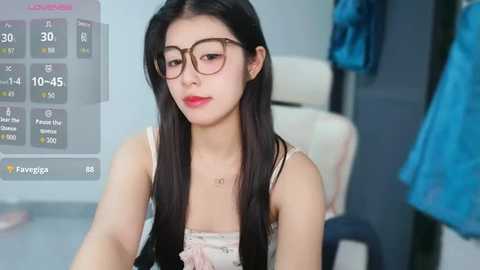 Video of an Asian woman with long black hair, wearing glasses and a white lace top, seated in a modern room with a blue garment hanging.