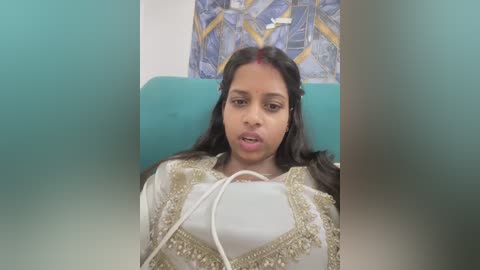 Video of a South Asian woman in a hospital bed, wearing a white saree with gold embroidery, red dot on her forehead, and a medical tube in her mouth.