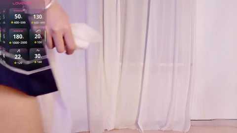 Video of a person with a light complexion holding a white cloth to clean a window, partially obscured by sheer curtains. The background displays a digital display with weather information.