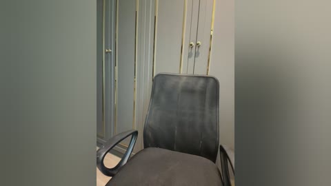 Video of a minimalist, modern office chair with a black leather-like seat, positioned against a muted green wall with vertical gold stripes and two small gold wall sconces. The chair's armrests and backrest are made of a textured black material.