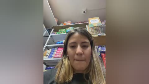 Video of a woman with long, straight, blonde hair, wearing a black dress, standing in a brightly lit, cluttered room with shelves full of colorful boxes and bags, including a large box of cereal.