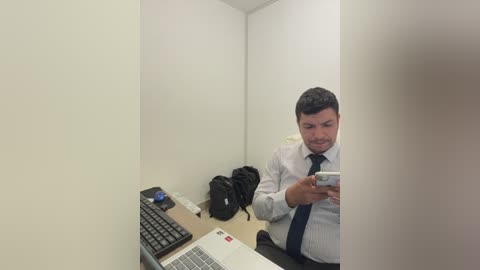 Video of a man with short dark hair, wearing a white shirt and dark tie, focused on a silver laptop. A black backpack and a cellphone are on a wooden desk in a small, white-walled cubicle.