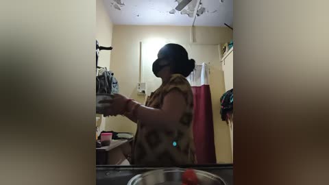 Video of a person in VR goggles, wearing a floral-patterned shirt, using a sink in a dimly-lit, cluttered bathroom with a red curtain and a white towel.