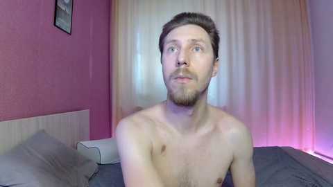 Video of a fair-skinned, bearded, shirtless man with short brown hair, gazing thoughtfully in a dimly lit bedroom with pink walls and a white curtain.