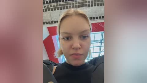 Video of a young Caucasian woman with blonde hair, wearing a black shirt, taken indoors. She appears serious, with a neutral expression. Background features red and white geometric patterns and a grid-patterned ceiling.