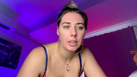 Video of a young Caucasian woman with light skin, brown hair tied in a bun, wearing a blue and white spaghetti strap top, looking serious in a dimly lit room with purple lighting.