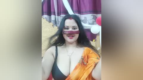 Video of a plus-size woman with long black hair, wearing a black bra, orange sari, and red makeup, sitting on a bed with a gold blanket, in a festive room with purple and white curtains.