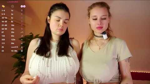 Video of two young women with large breasts, one in a white sweater, the other in a beige top, indoors, warm lighting.