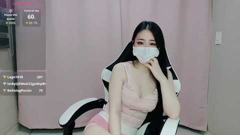 A video of an East Asian woman with long black hair, wearing a pink camisole and pink shorts, sitting in a black and white gaming chair. She has a surgical mask covering her mouth and nose.