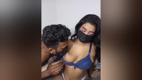 Video: A dark-skinned man with curly hair, wearing a mask, leans in to kiss a light-skinned woman with long black hair and blue bra, who's also masked, in a dimly lit room.