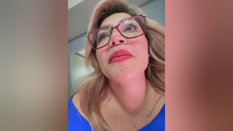 A video of a blonde, middle-aged woman with glasses, red lipstick, and a blue top, taken from a low angle.
