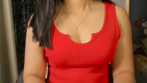 Video of a woman with medium skin tone and straight black hair, wearing a tight, sleeveless red top, showcasing medium-sized breasts. She has a delicate necklace with a small pendant. The background is blurred, featuring a dark curtain and indistinct objects.