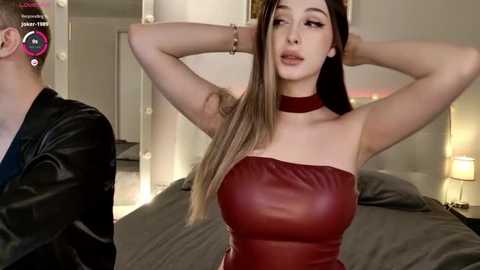 Video of a fair-skinned woman with long brown hair, wearing a tight, red leather strapless dress, posing in a modern bedroom.