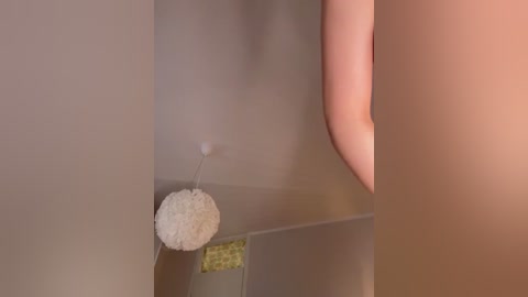 A video of a beige wall with a white, fluffy pom-pom attached to it by a thin string, slightly hanging, and a blurred, partially visible person on the right.