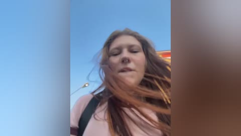 Video of a Caucasian woman with long, reddish-brown hair, wearing a pink top, standing against a clear blue sky. The image is slightly out of focus, capturing a candid moment with a playful, windblown effect.