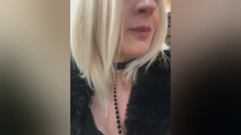 A close-up video of a woman with straight, platinum blonde hair and fair skin, wearing a black, textured sweater with a black choker necklace. Her lips are slightly parted, and she appears to be smiling. The background is blurred.