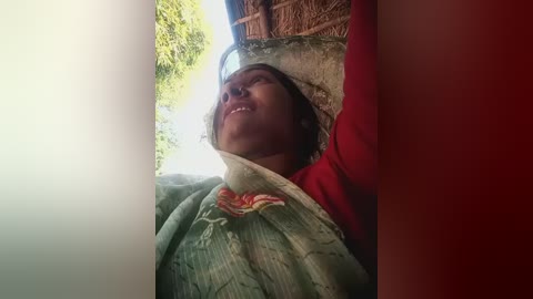 Video of a woman with medium brown skin and short dark hair, wearing a red shirt, lying under a green floral-patterned blanket, with a thatched roof and lush greenery in the background.