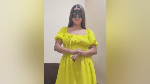 Media: Video of a young woman with long dark hair, wearing a bright yellow dress, black mask, and holding hands, standing in a room with yellow and brown walls.