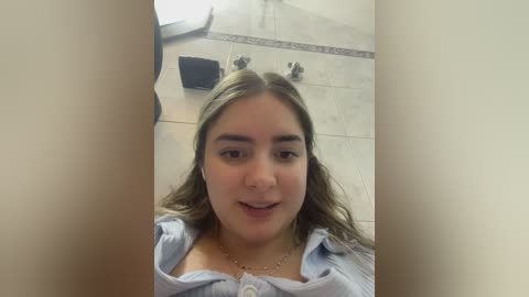 Video of a young woman with light skin and brown hair, wearing a light blue shirt, standing in a tiled bathroom with beige walls.