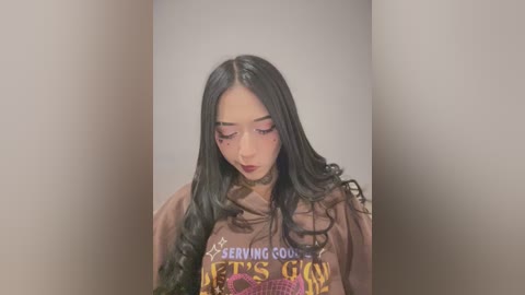 Video of a young East Asian woman with long, black, wavy hair, wearing a brown hoodie with \"SERVING GOD\" in yellow text, standing against a plain beige wall, eyes closed, expression contemplative.