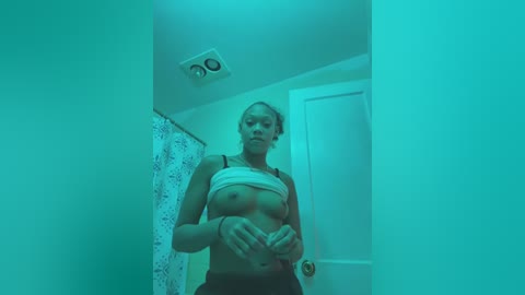 Video of a topless woman with fair skin and blonde hair, wearing a white crop top and black shorts, in a dimly lit bathroom with a greenish tint.