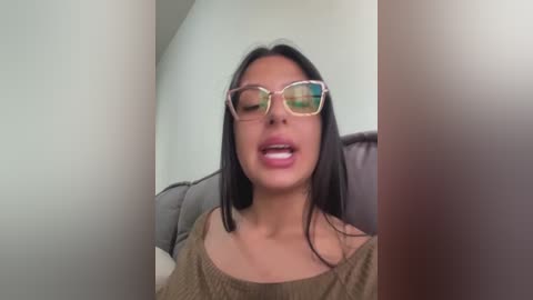 Video of a woman with straight black hair, wearing glasses, a beige off-shoulder top, and sitting on a brown couch against a plain white wall.