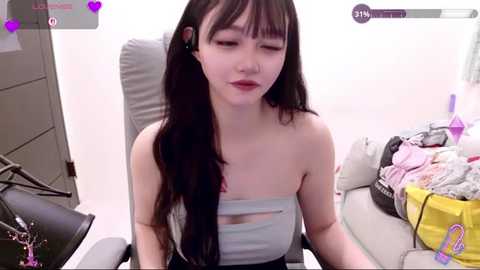 Video of an Asian woman with long black hair and fair skin, wearing a strapless light blue top, sitting in an office chair amidst a cluttered desk with clothes, bags, and a laptop.