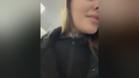 Video of a woman with light skin, brown hair, and wearing a black jacket, smiling slightly. Background is blurry, suggesting an indoor setting.