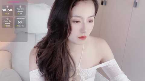 Media: A video of an East Asian woman with long black hair, fair skin, and red lipstick, wearing an off-shoulder white lace top, standing in a softly lit room with a white bed and blurred background.