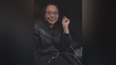 Video of a smiling woman with curly hair, wearing glasses and a black jacket, seated in a dimly-lit room, holding a microphone.