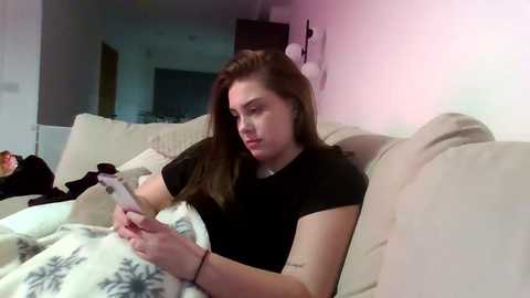 Video of a young woman with light skin and long brown hair, wearing a black shirt, lying on a beige couch, holding a smartphone, in a dimly lit living room.