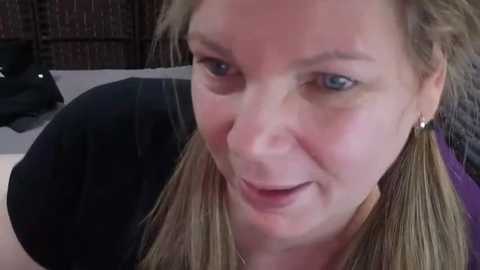 A close-up video of a light-skinned, smiling woman with blonde hair tied back, wearing a black shirt and earrings. Background shows a blurred, dark wooden frame.