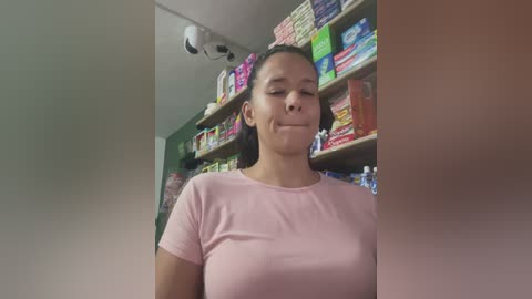 Video of a young woman with a light complexion and dark hair tied back, wearing a pink t-shirt, smiling in a brightly lit, colorful candy store with shelves filled with various candies.